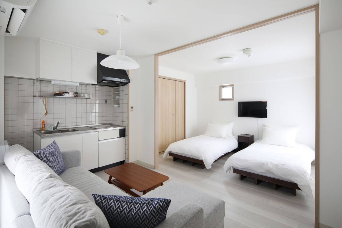 A modern hotel room with a kitchenette, a sofa, two beds, and a wooden coffee table.