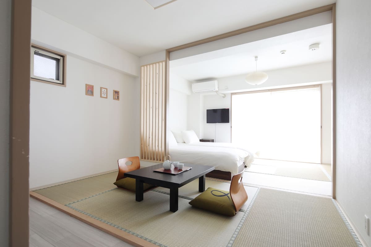 A minimalist hotel room with tatami flooring, a bed, a small table with tea set, and a sliding door leading to a balcony.