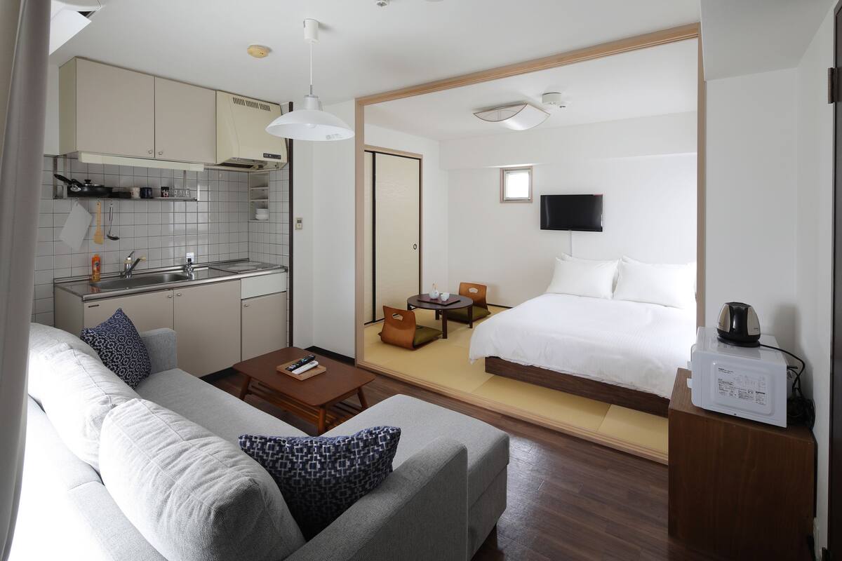 A compact hotel room with a kitchenette, a sofa, a bed, and a TV.