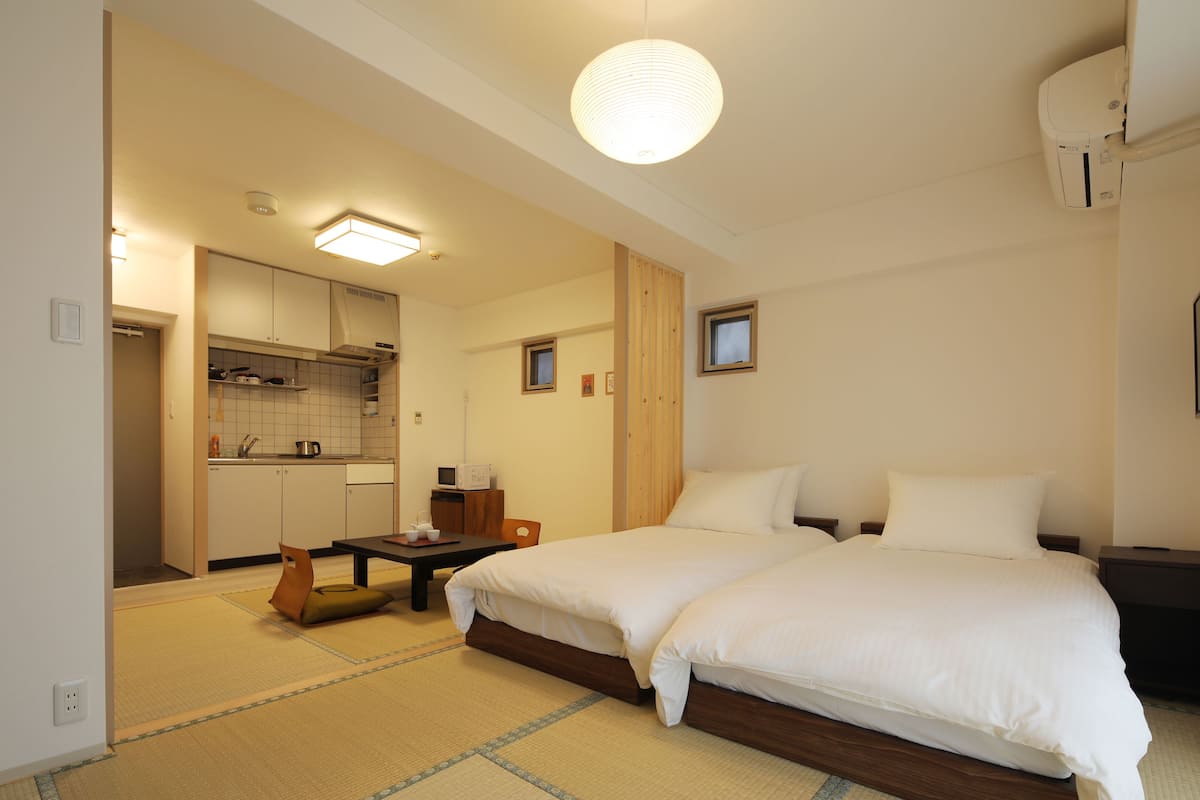 A hotel room with a bed, a small table, a chair, a kitchen area, and a ceiling light.
