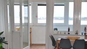 Une pièce comprenant une table à manger, des chaises, une plante et offrant une vue sur une étendue d’eau grâce à de grandes fenêtres.