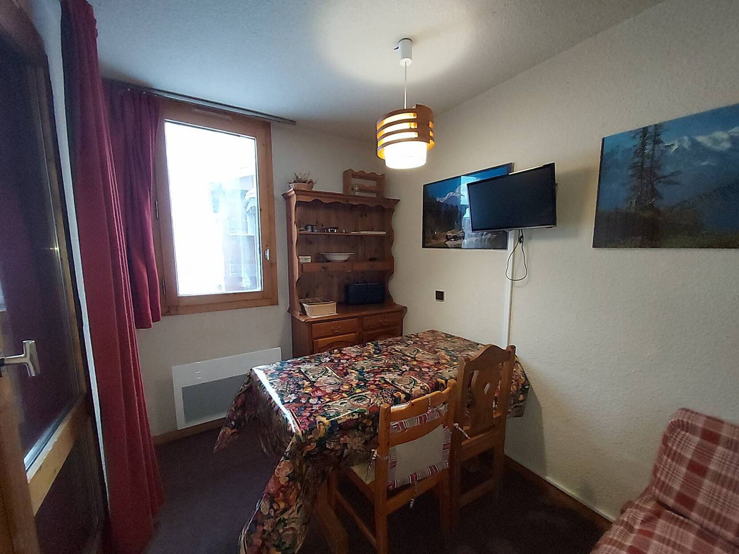 Ein kleiner Raum mit einem Holztisch, der mit einer Blumen Tischdecke bedeckt ist, einem an der Wand befestigten Fernseher und einem Fenster mit roten Vorhängen.