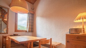 Une table à manger en bois avec des chaises, un luminaire suspendu, une fenêtre avec des rideaux et un meuble en bois contenant une radio.
