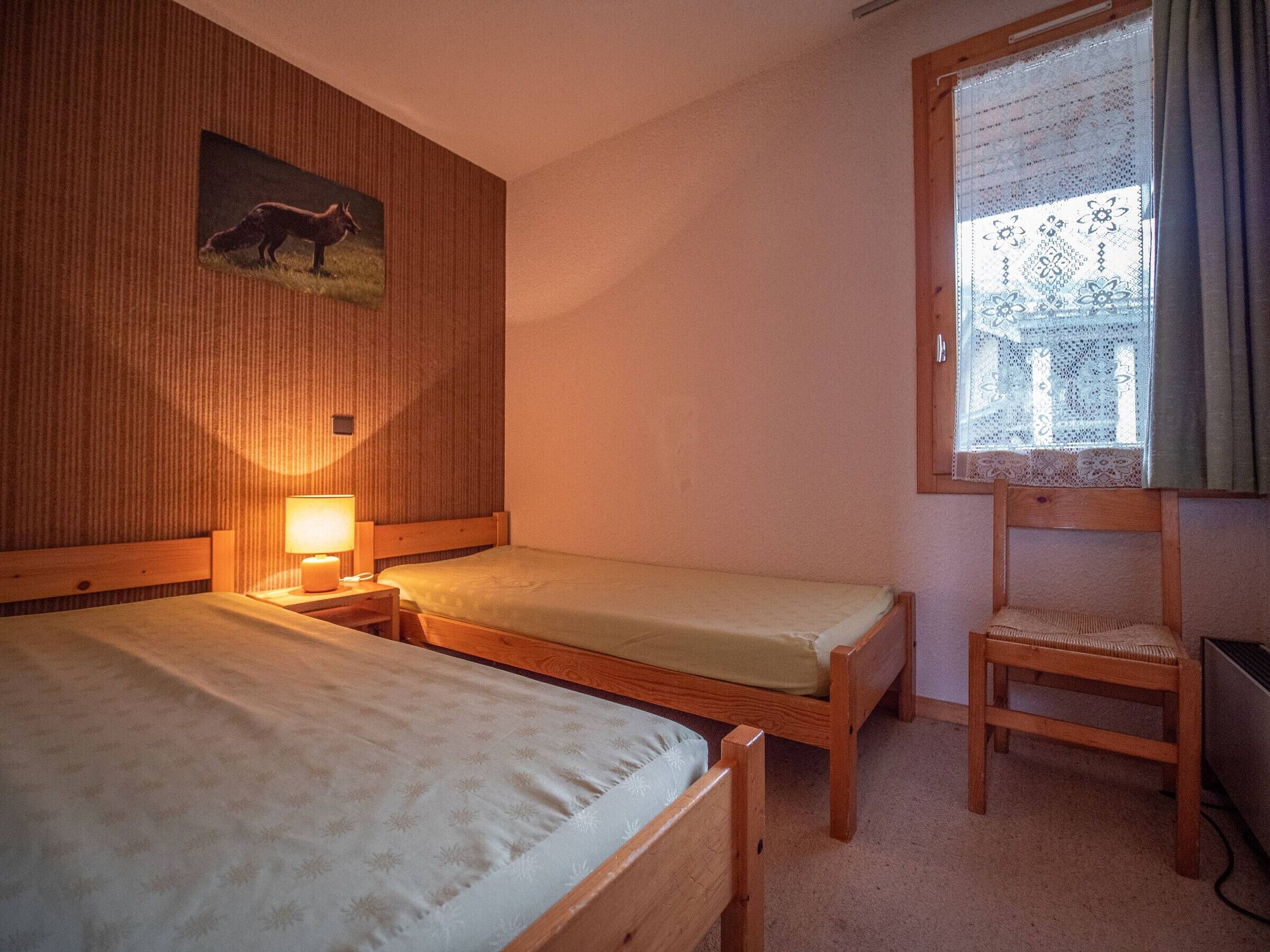 A small, cozy room with two wooden beds, a nightstand with a lamp, a wooden chair, and a window with lace curtains.