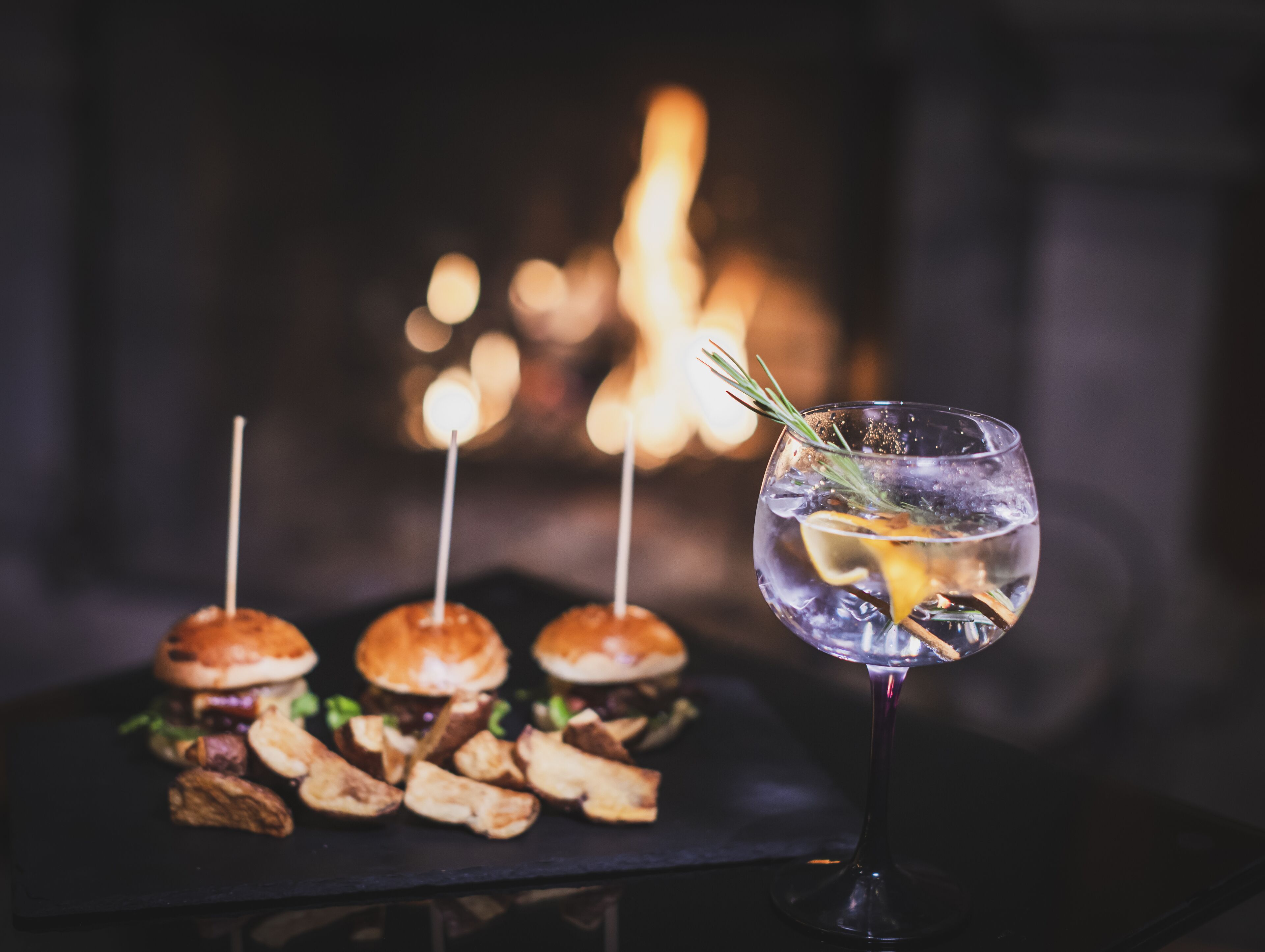 Una mesa con tres hamburguesas y un vaso de bebida transparente, adornada, sobre un fondo desenfocado con iluminación cálida.