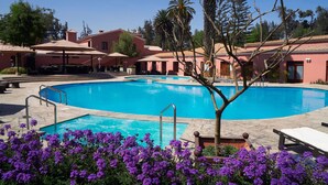 Une piscine à l’eau d’un bleu limpide, entourée d’un bâtiment à la façade rose, et un arbre au premier plan.