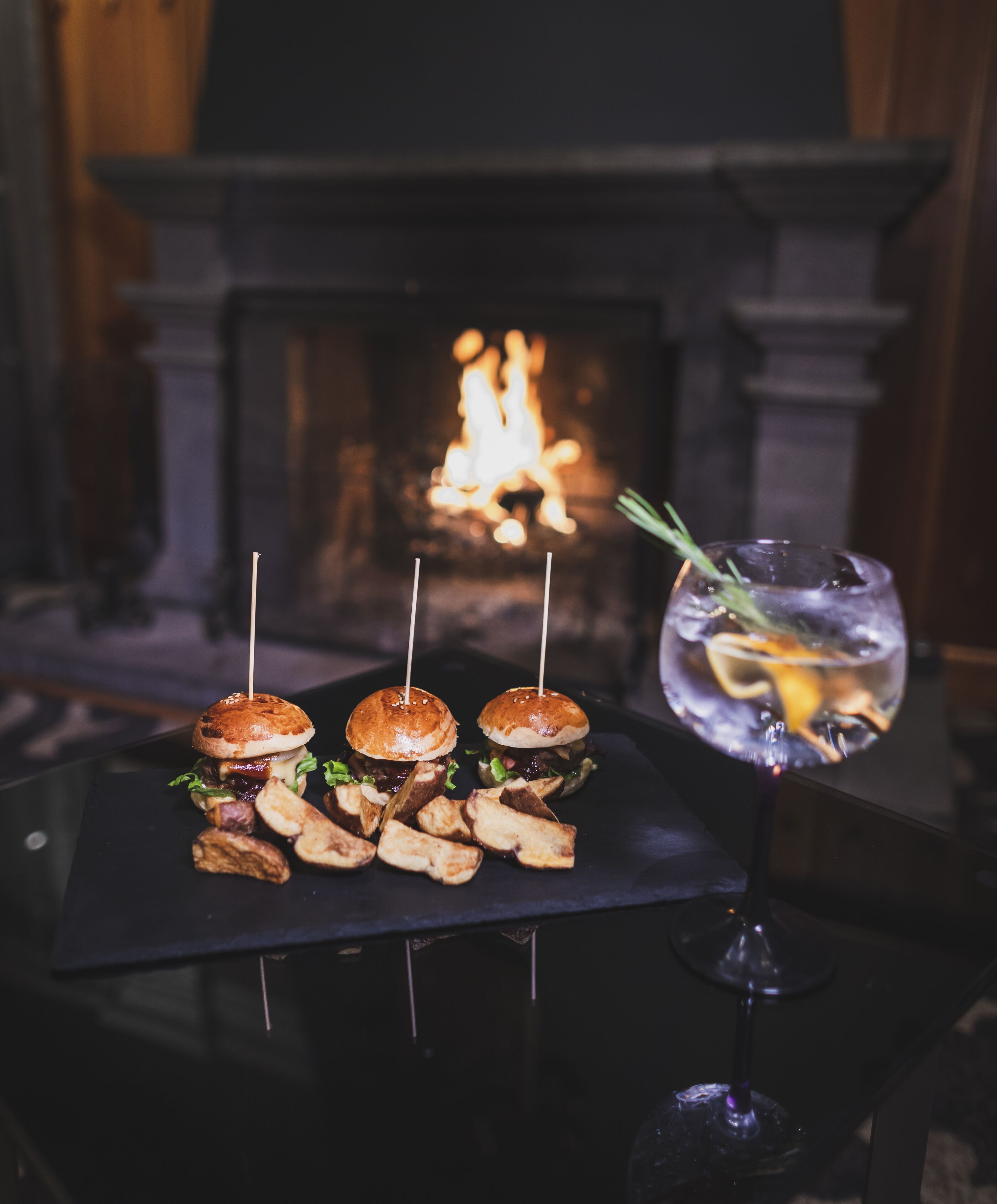 A table with three burgers and a glass of iced drink, set against a background of a fireplace.