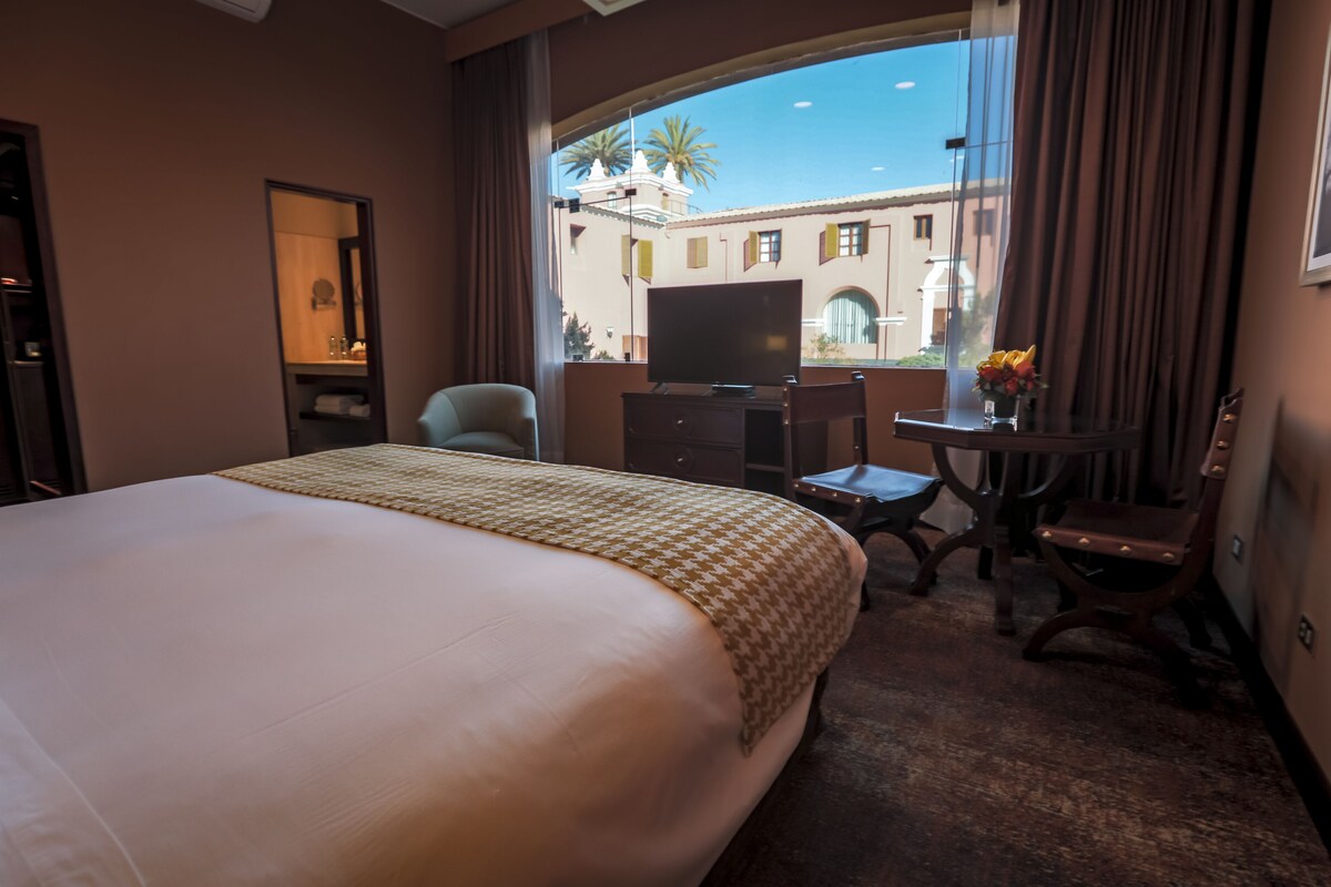 Une chambre d’hôtel avec un grand lit, un bureau avec une chaise, une télévision et une fenêtre donnant sur un immeuble et des palmiers.