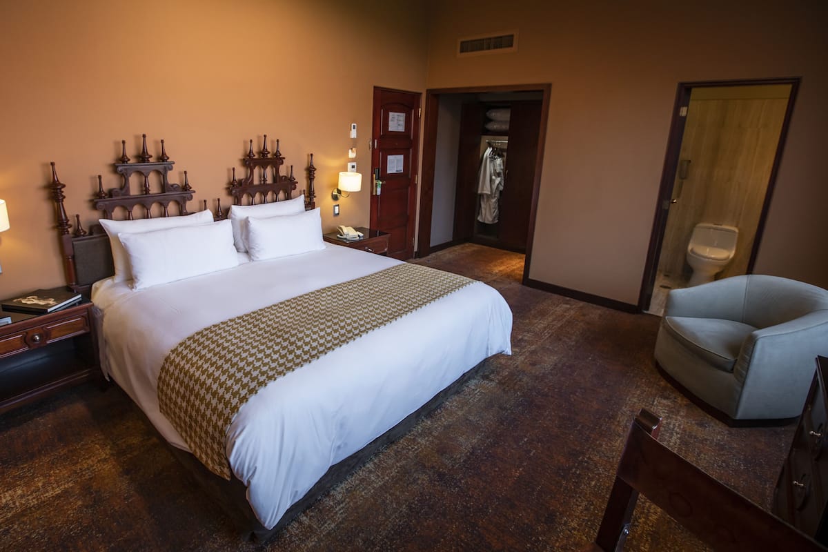 Une chambre d’hôtel avec un grand lit, des lampes de chevet, une tête de lit en bois, une armoire, une salle de bain avec des toilettes et une chaise.