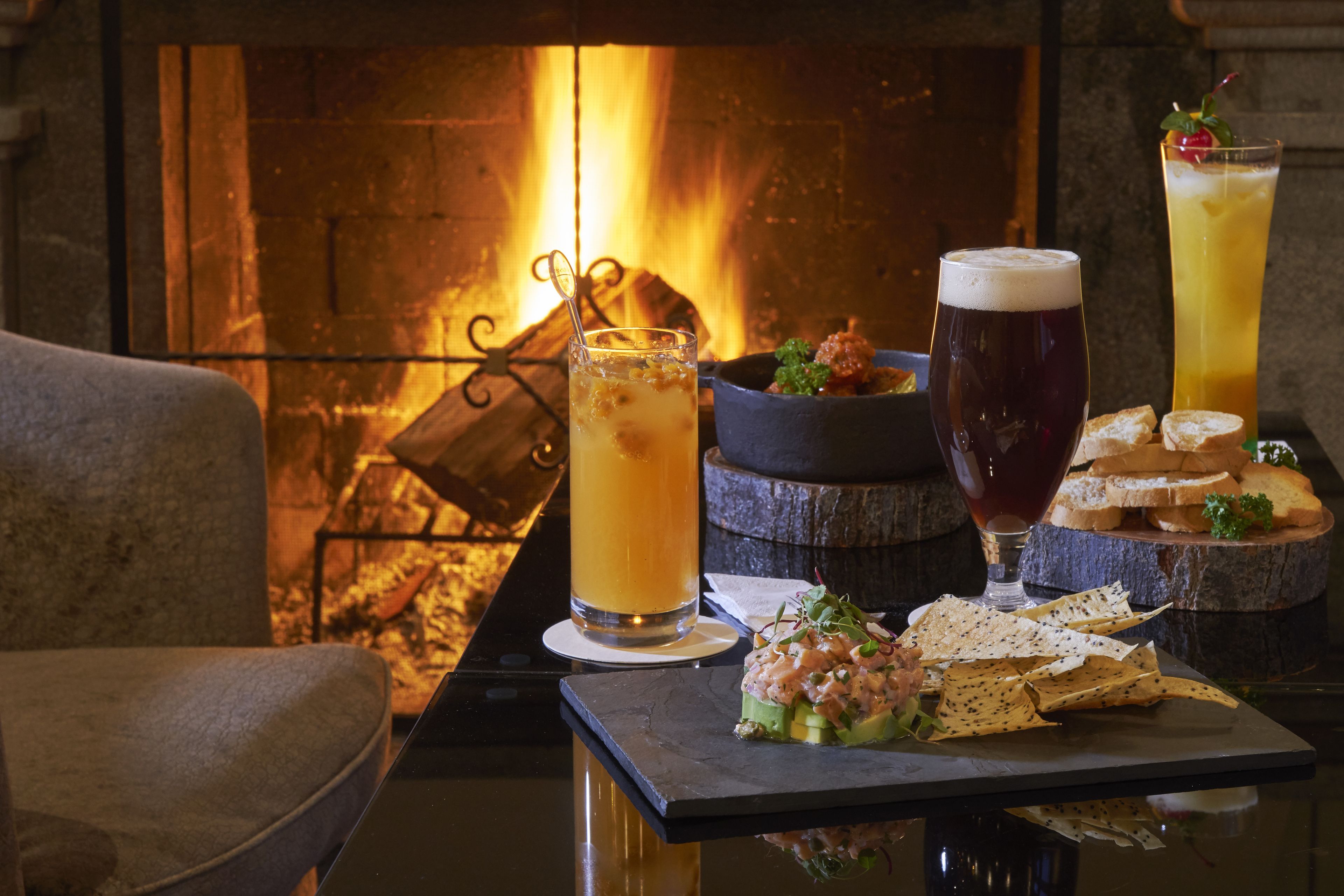 A table with a glass of beer, a tall glass of a yellow beverage, and a bowl of food, set against a fireplace backdrop.
