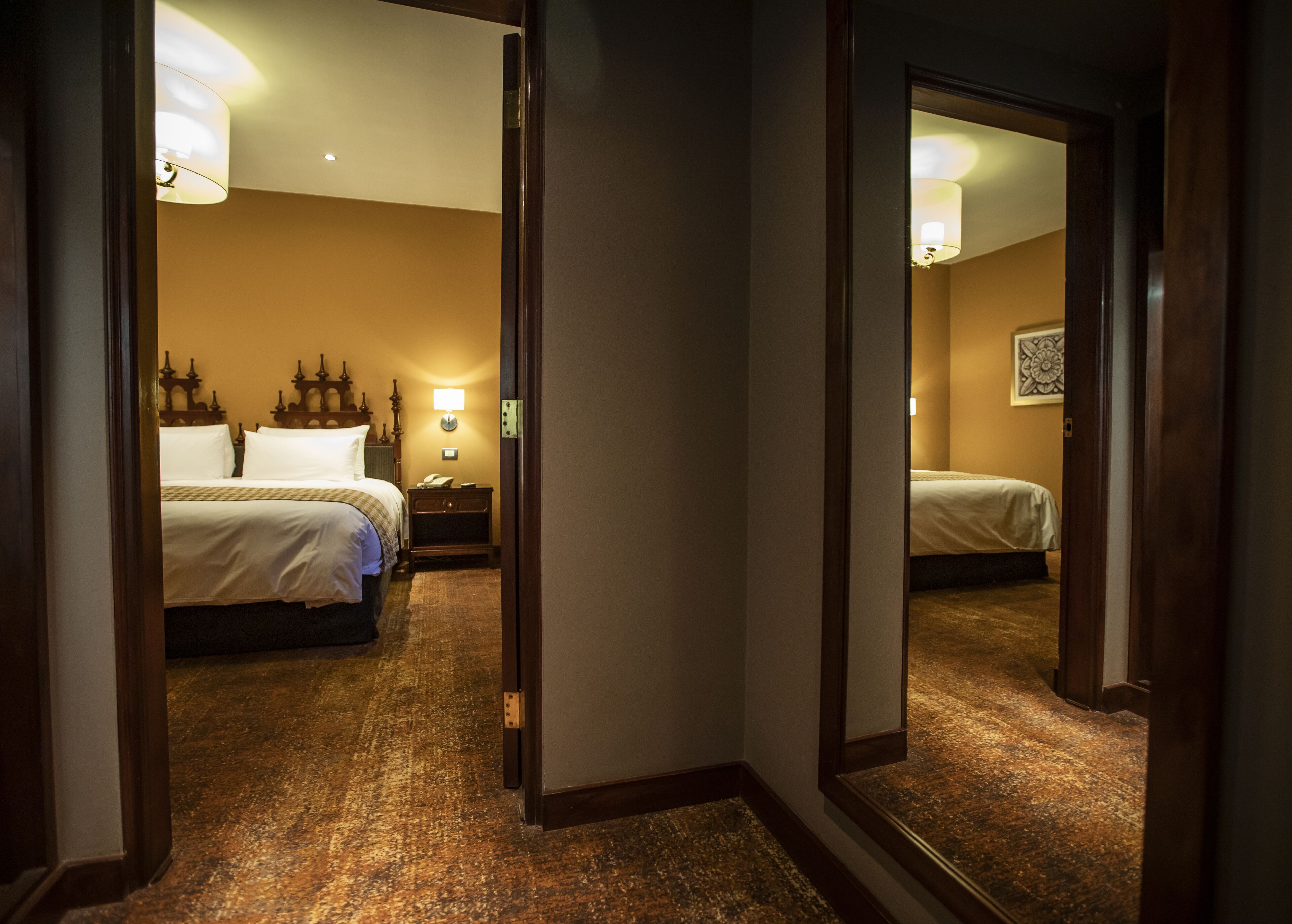 A hotel room with a large bed, bedside table, lamp, and a mirror reflecting the room.