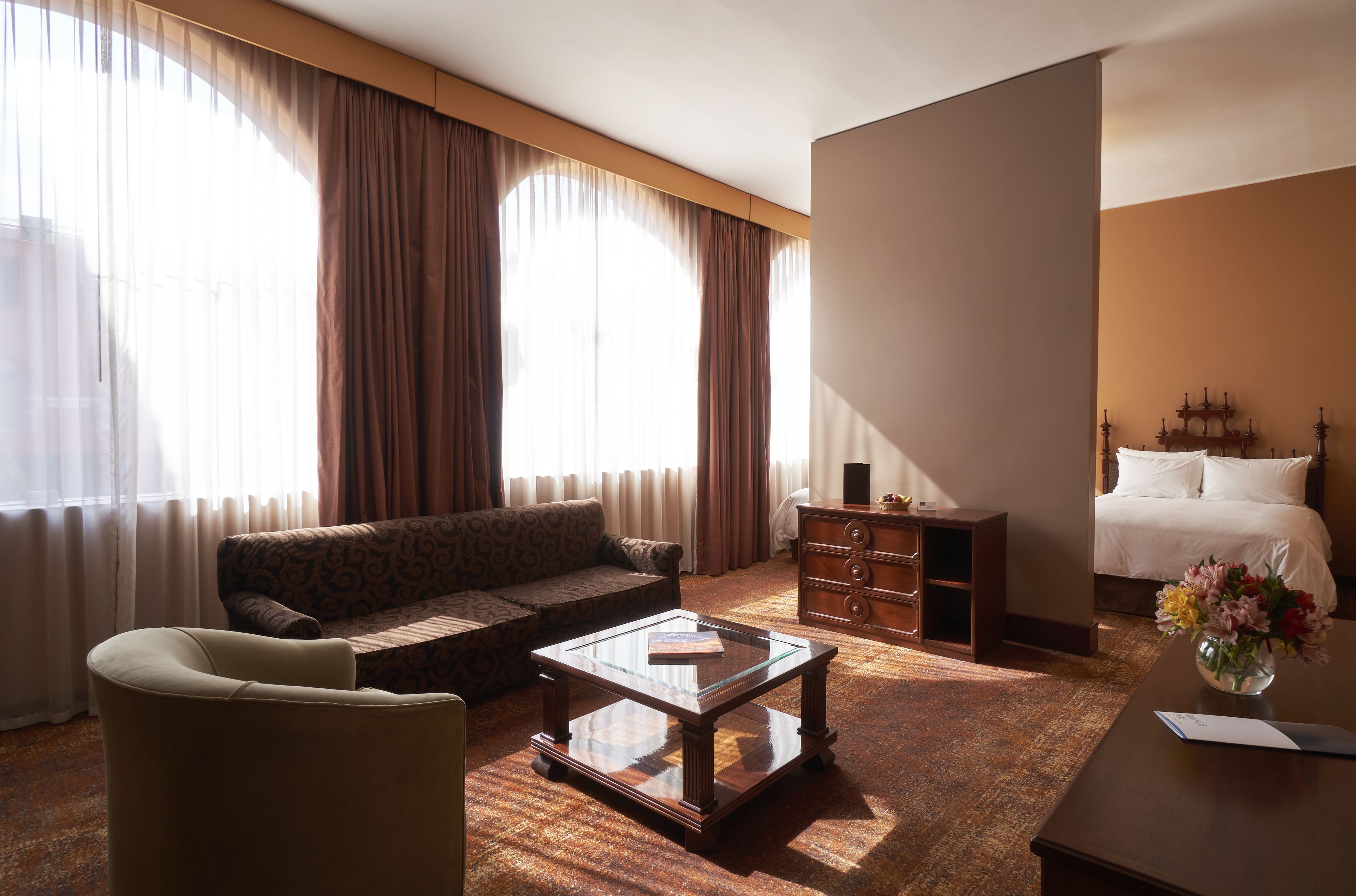 Habitación de hotel con sofá, una mesa de centro de vidrio, cómoda de madera y cama con ropa blanca.