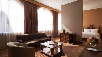 Habitación de hotel con sofá, una mesa de centro de vidrio, cómoda de madera y cama con ropa blanca.
