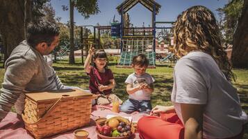 Un picnic familiar con una cesta, frutas y una botella de jugo sobre una manta en un parque.