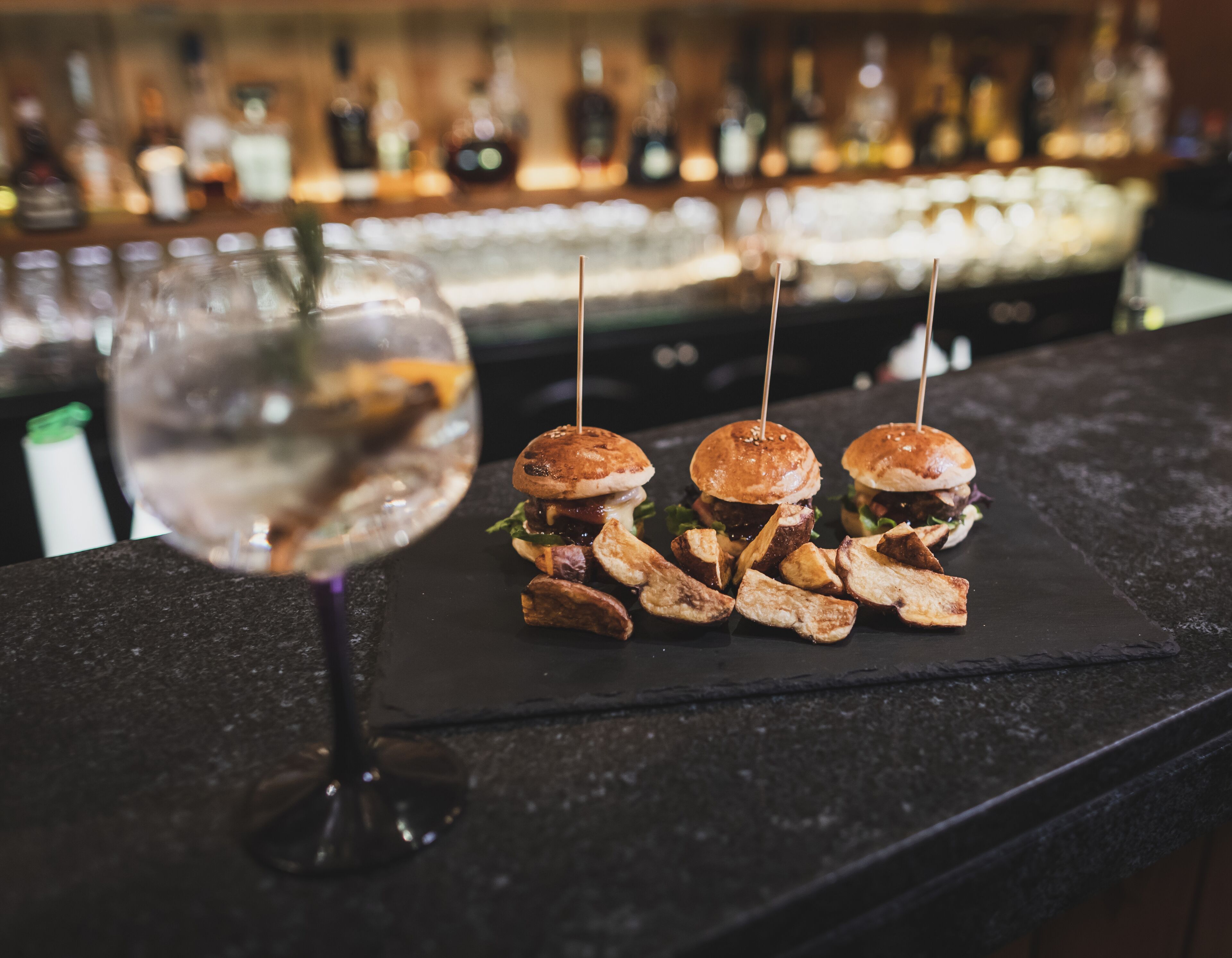 A bar setting with a glass of water and three burgers with toothpicks on a black slate.