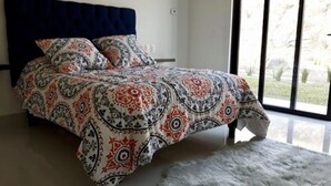 Een slaapkamer met een bed, een blauw hoofdeinde en uitzicht op het omliggende landschap door grote ramen.