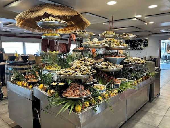 A buffet with various seafood dishes, including oysters and shrimp, arranged on metal trays and surrounded by tropical plants. - Pavillon T3 *** 72m2 Terrace 180° Sea View, Wifi, Swimming pool, Internet, Air conditioning, (Fleury)