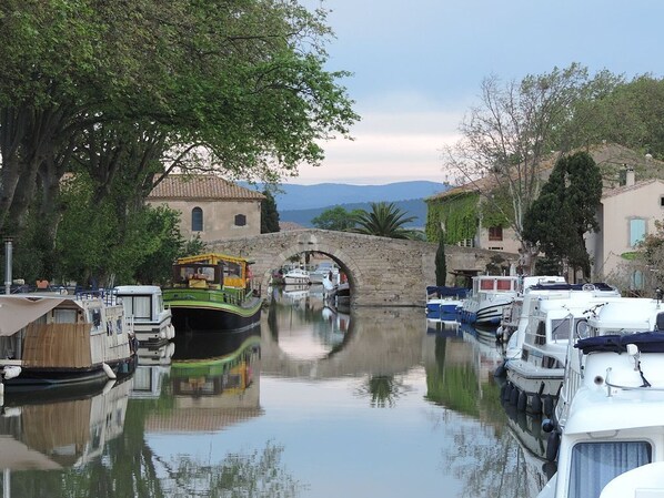 Un canal où sont amarrés des bateaux, un pont de pierre et des bâtiments bordés d’arbres.