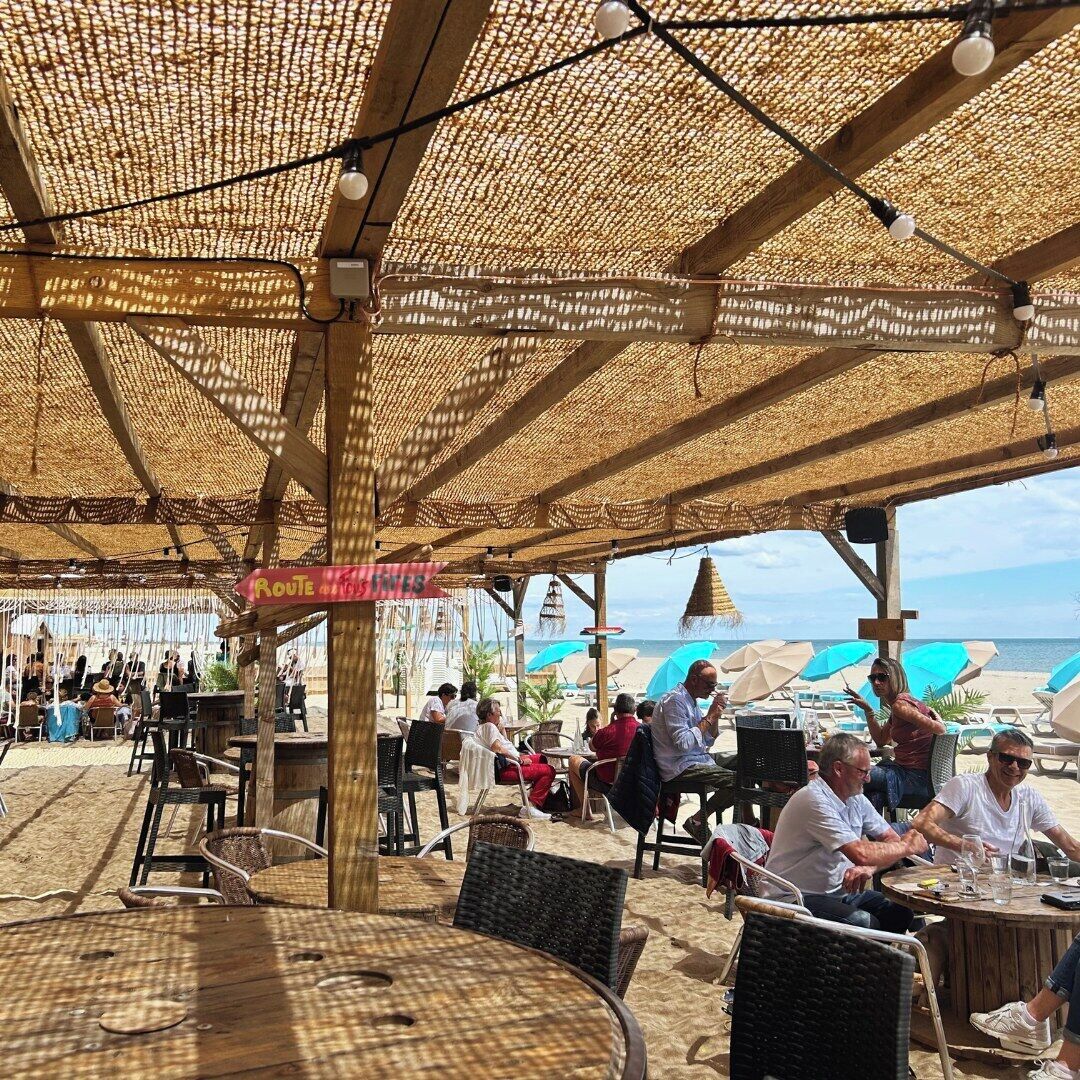 Ein Außenbereich mit Tischen und Stühlen unter einem Strohdach mit Blick auf einen Strand.