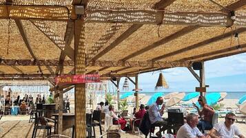 Ein Außenbereich mit Tischen und Stühlen unter einem Strohdach mit Blick auf einen Strand.