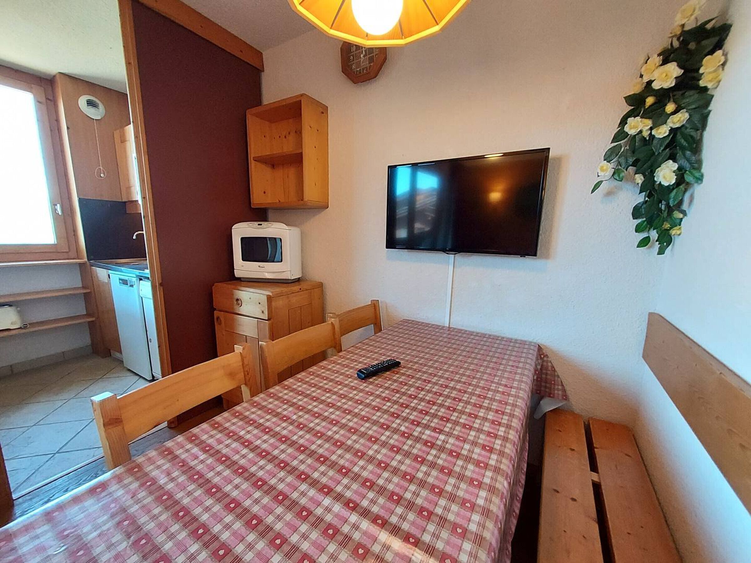 Ein kleines Zimmer mit Küchenzeile, einem Bett mit kariertem Überwurf, einem an der Wand befestigten Fernseher und einer kleinen Holzkabine.