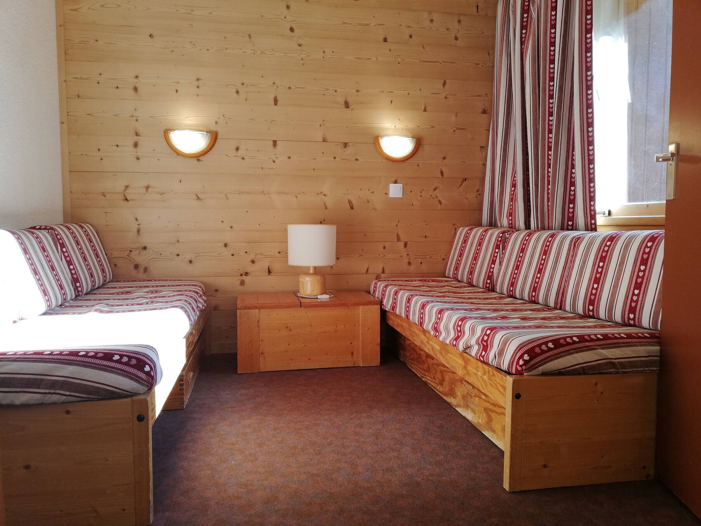 Una habitación acogedora con dos camas de madera, una mesita de noche de madera con lámpara, y una ventana con cortinas.