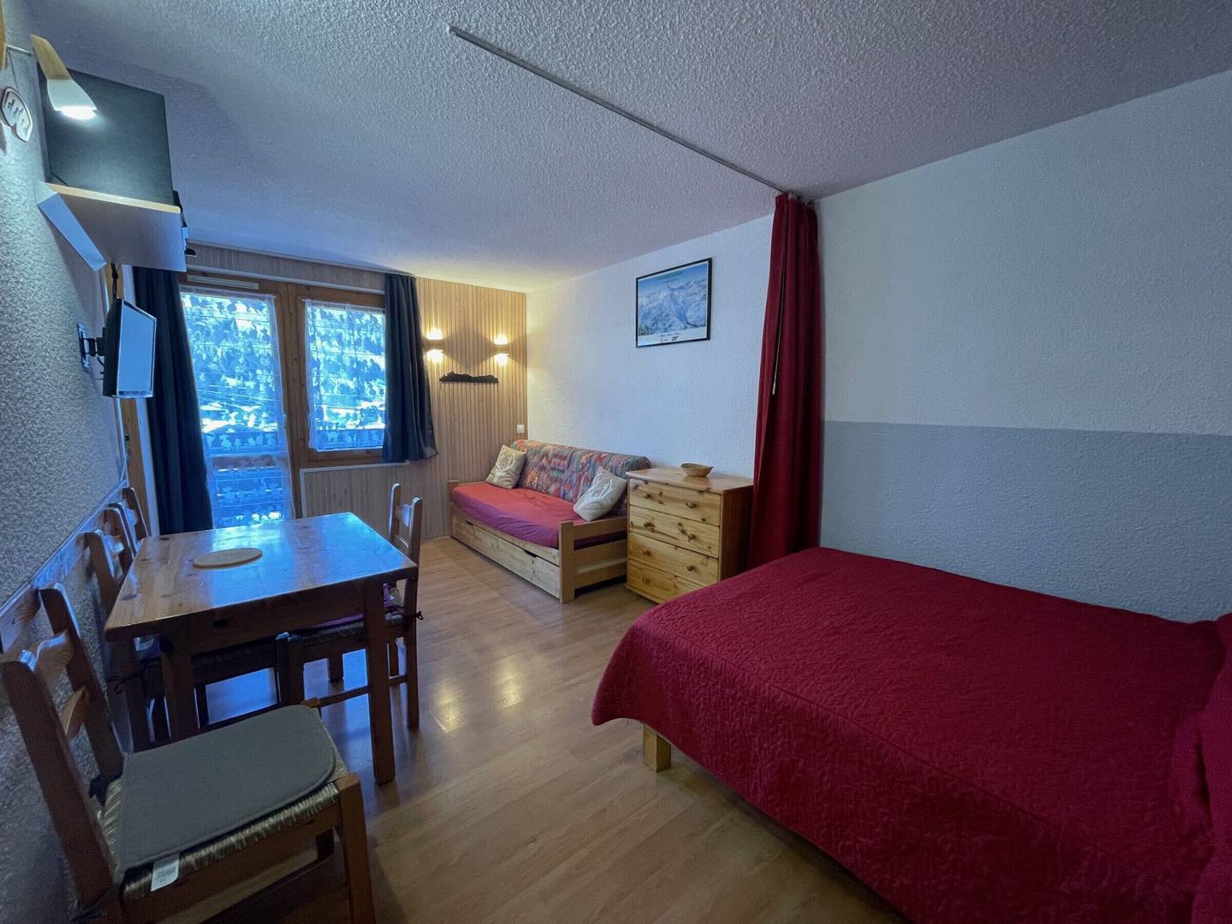 Una habitación funcional con cama, sofá, mesa de comedor con sillas, televisor montado en la pared y ventana con cortinas.