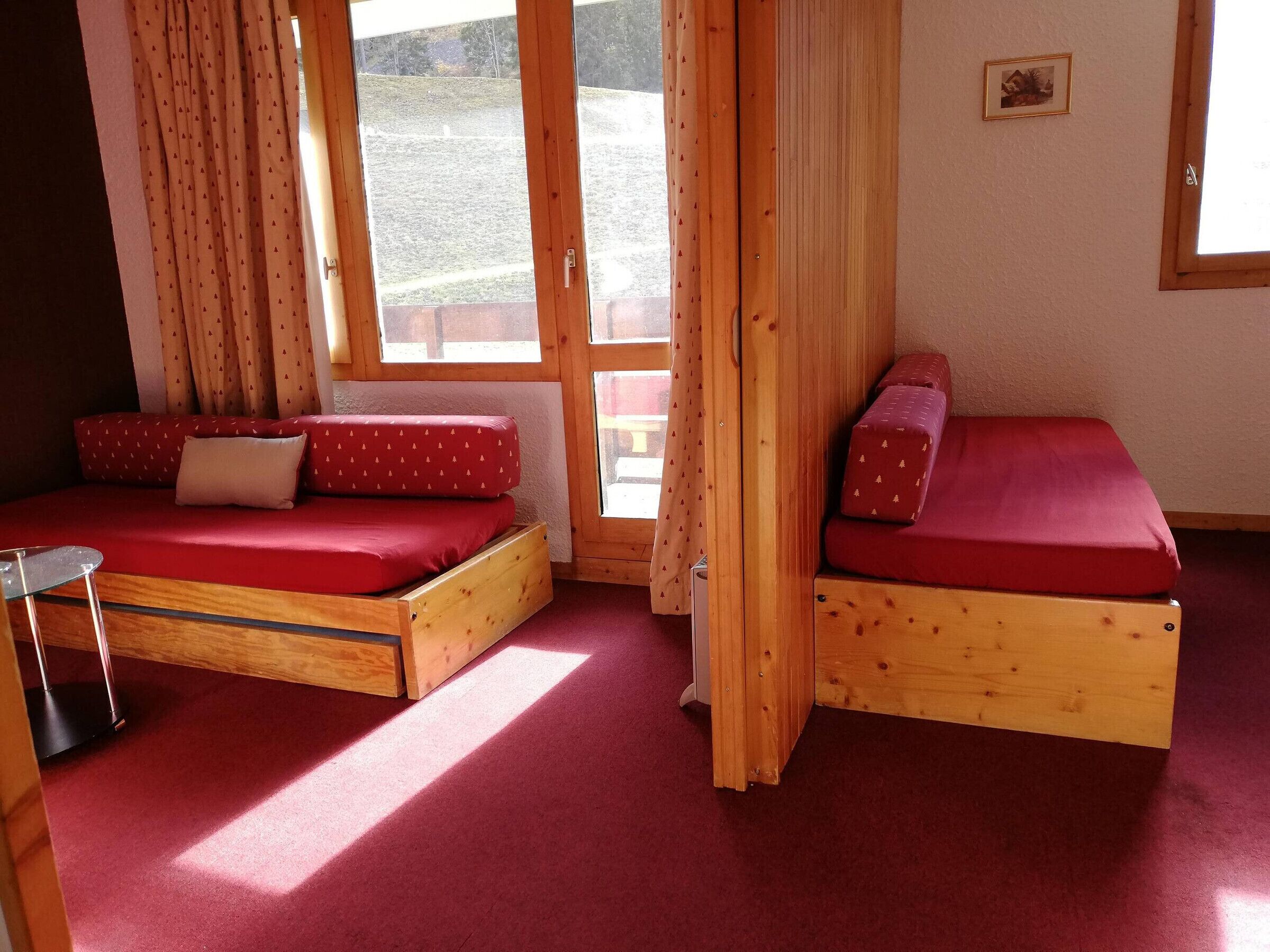 A room with two wooden beds with red bedding, a small table, and large windows with curtains.