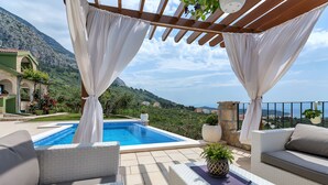 Zona junto a la piscina con mobiliario de mimbre blanco, vista al mar y paisaje montañoso.