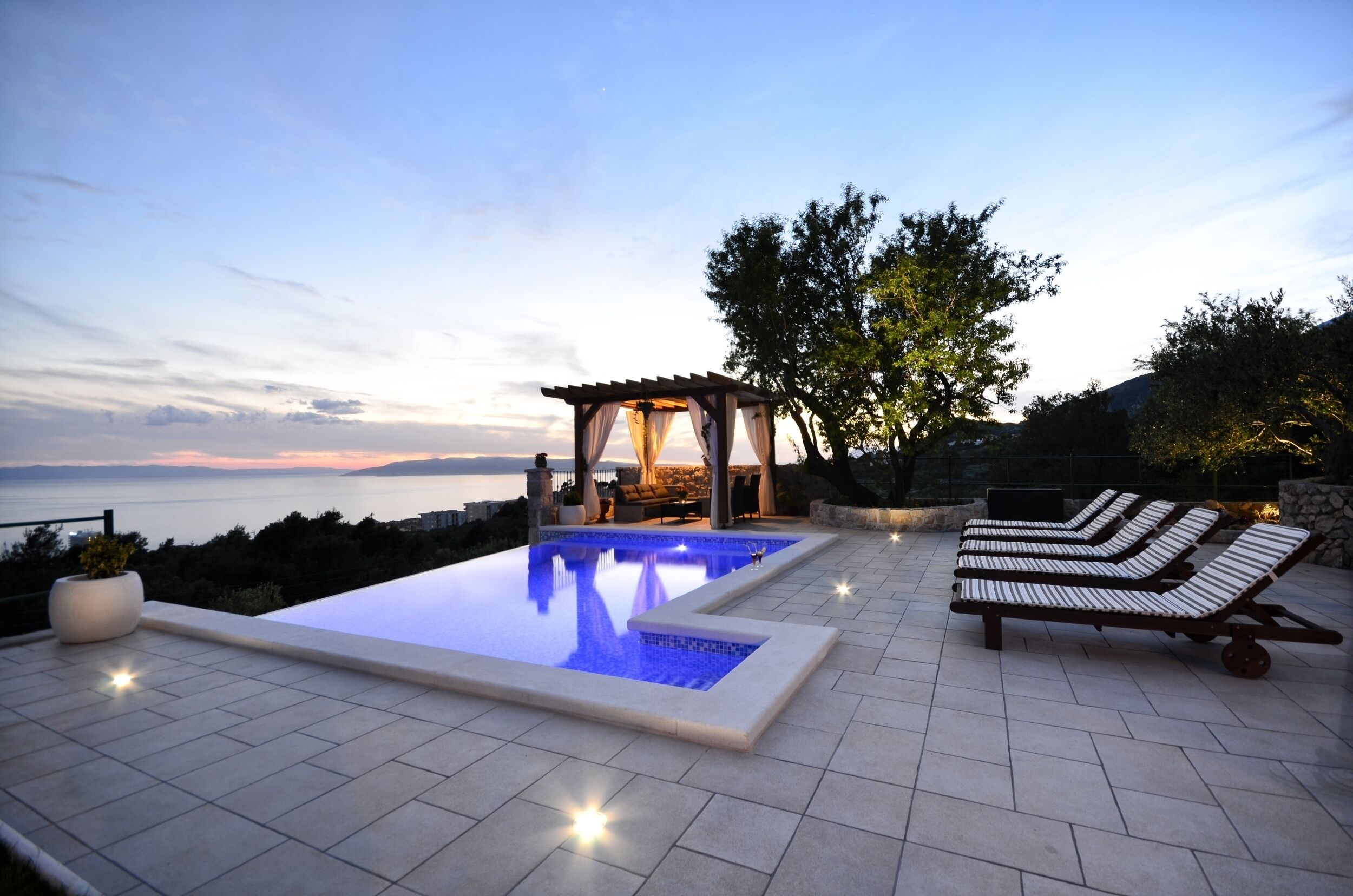 A rooftop infinity pool with a pergola and lounge chairs, overlooking a scenic view.