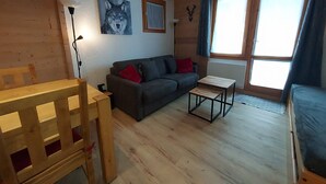 A cozy living room with a wooden floor, a grey sofa, a small wooden table, and a window with curtains. - 2-room apartment in Valmorel, near slopes and shops, equipped, wifi, non-smoking (Valmorel)