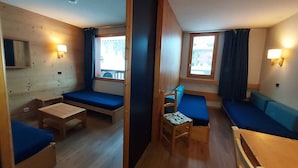 A compact room with a bed, a small table, a chair, and a sofa. There are two windows with curtains, a wall-mounted lamp, and a wooden floor.