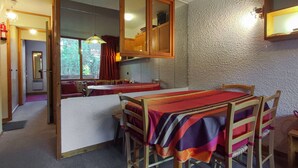 Un comedor con mesa y sillas de madera, una zona de cocina con armarios, y vistas a un bosque por la ventana.