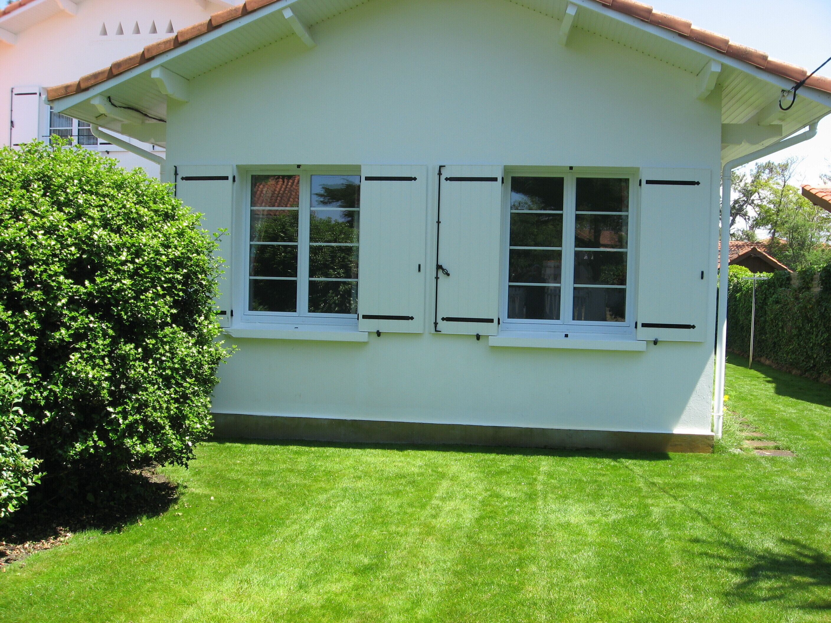 A white house with two windows and a door, surrounded by a well-maintained lawn and a hedge.
