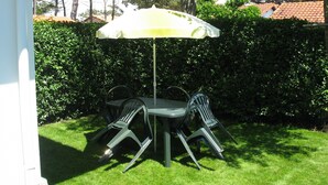 Een terras buiten met een tafel, stoelen, een parasol en een haag op de achtergrond.