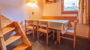 A cozy dining area with a wooden table and chairs, a small shelf with decorative items, and a window with curtains. - Divisible studio + mezzanine, ski-in/ski-out, sleeps 5, Valmorel (Valmorel)