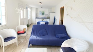Un salon chaleureux avec un canapé bleu, des fauteuils en osier blancs, une petite table et, au fond, une cuisine.