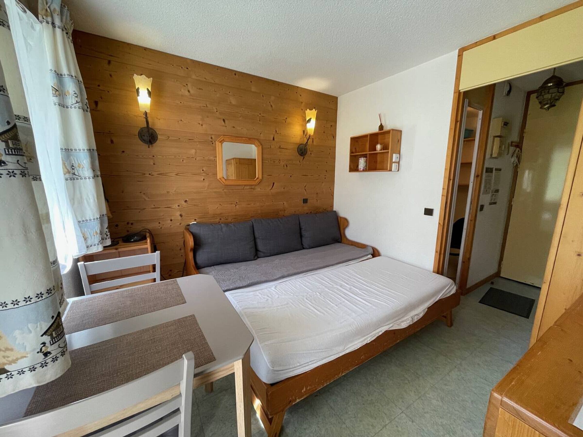 A bedroom with a wooden headboard, a bed with white bedding, a bedside table, a chair, and a mirror on the wall.