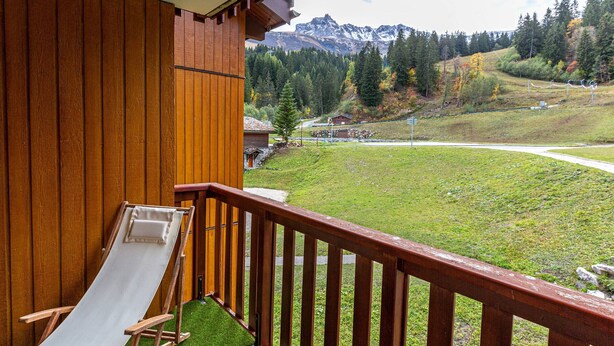 Un balcón de madera con una silla reclinable blanca, con vista a un paisaje montañoso y una perspectiva despejada de las montañas y una carretera sinuosa.