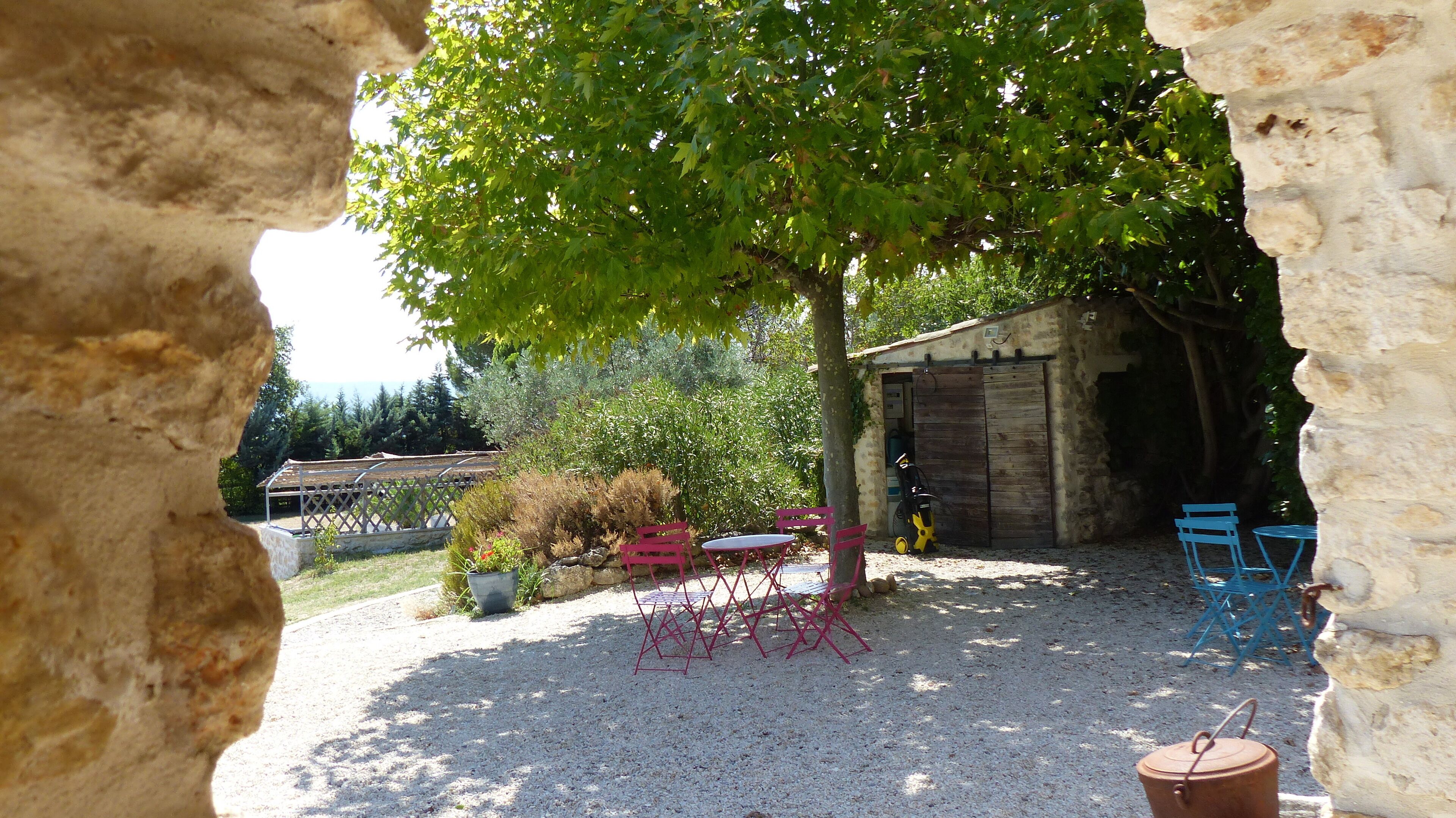 Un espace extérieur rustique aménagé avec un sol de gravier, un arbre qui offre de l’ombre et une structure en bois en arrière-plan.