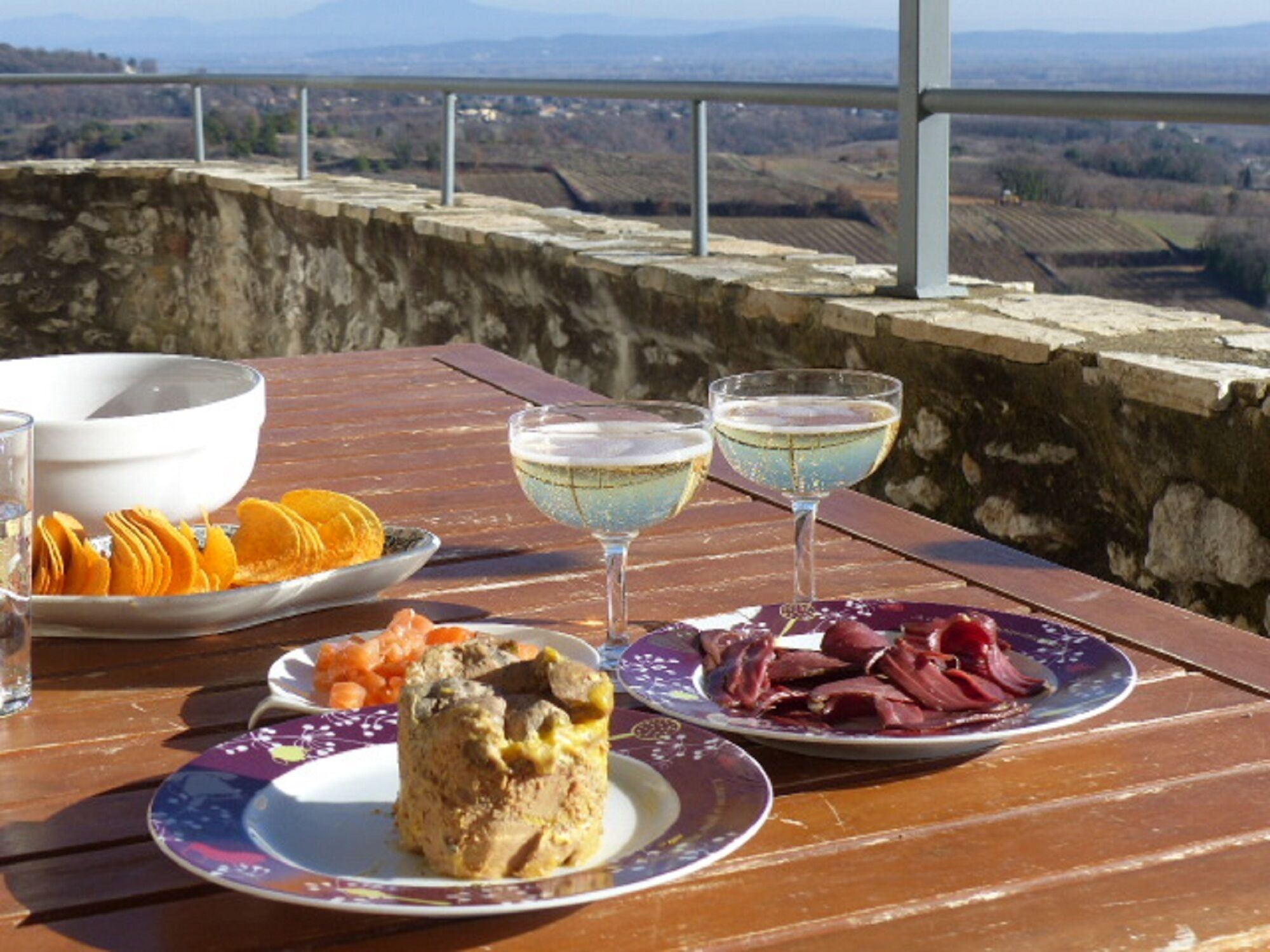 A table with a meal consisting of a cake, fruit, and meat, accompanied by two glasses of a light-colored beverage, set against a scenic view.