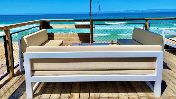 A wooden deck with a white cushioned bench under a beige umbrella, overlooking a beach with clear blue water and a wooden railing.