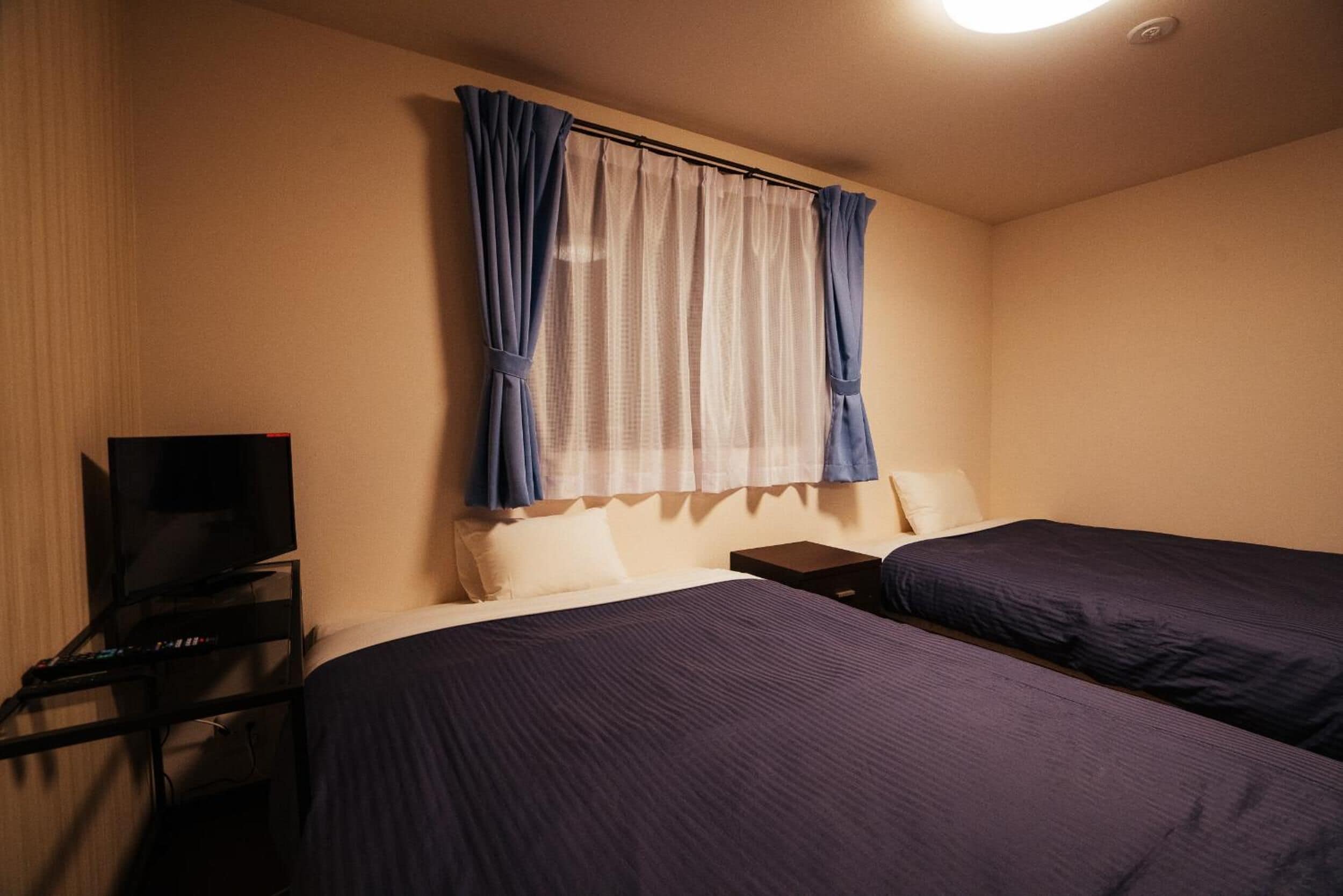 Une chambre d’hôtel avec deux lits, un téléviseur posé sur un meuble et une fenêtre avec des rideaux.