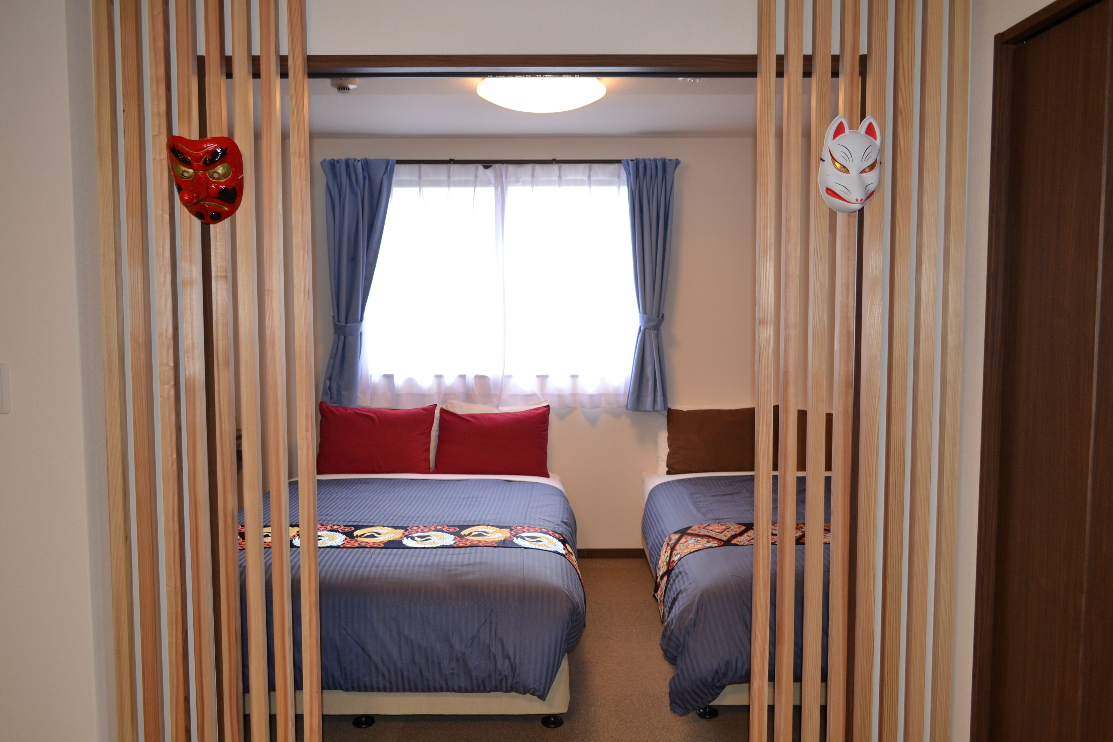 A room with two beds, a window with blue curtains, and a wooden partition with masks.