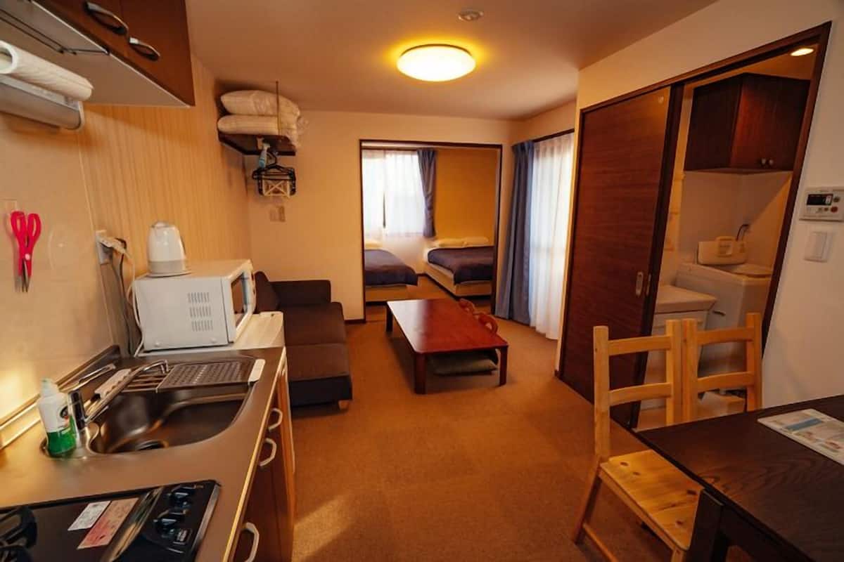 A compact hotel room with a kitchenette, a single bed, a sofa, and a wooden dining table.