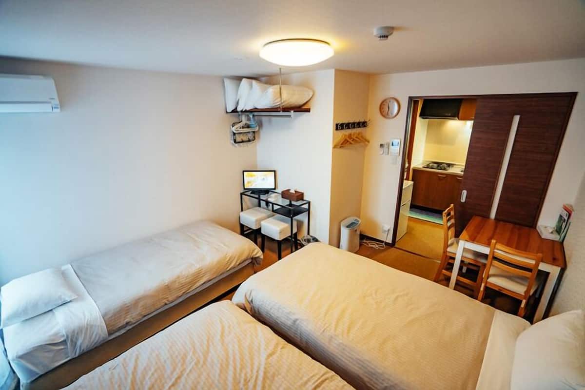 A compact hotel room with two single beds, a small desk with a computer, a chair, and a kitchenette.