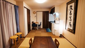 Een compacte hotelkamer met een eettafel, stoelen, een kleine keukenhoek en een traditioneel Japanse kunstwerk aan de muur.