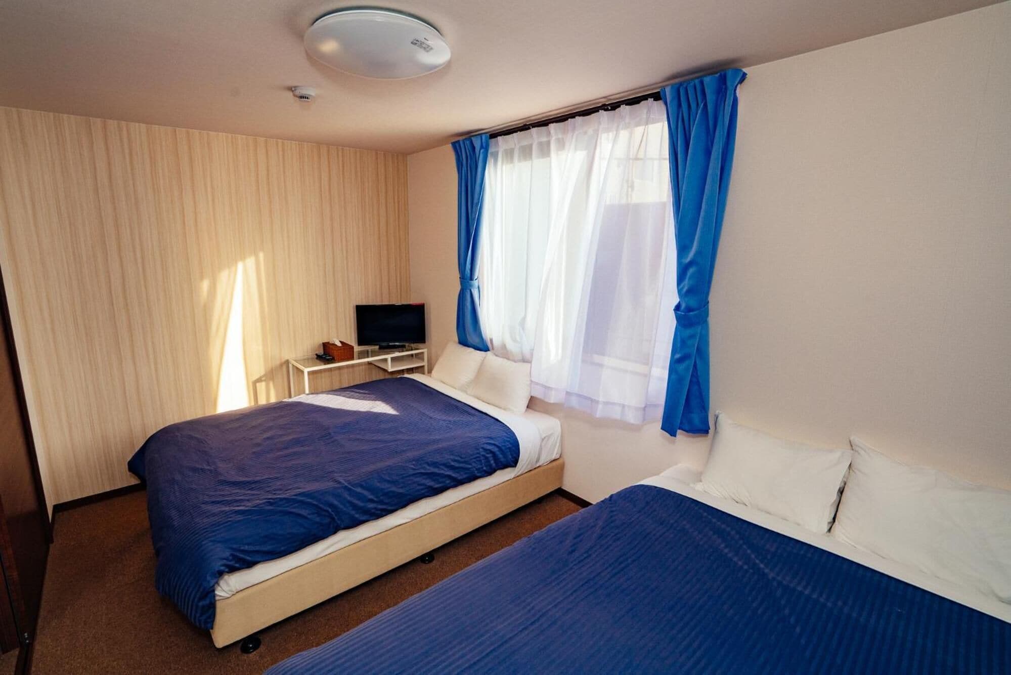 A hotel room with two beds, a television on a side table, and a window with blue curtains.