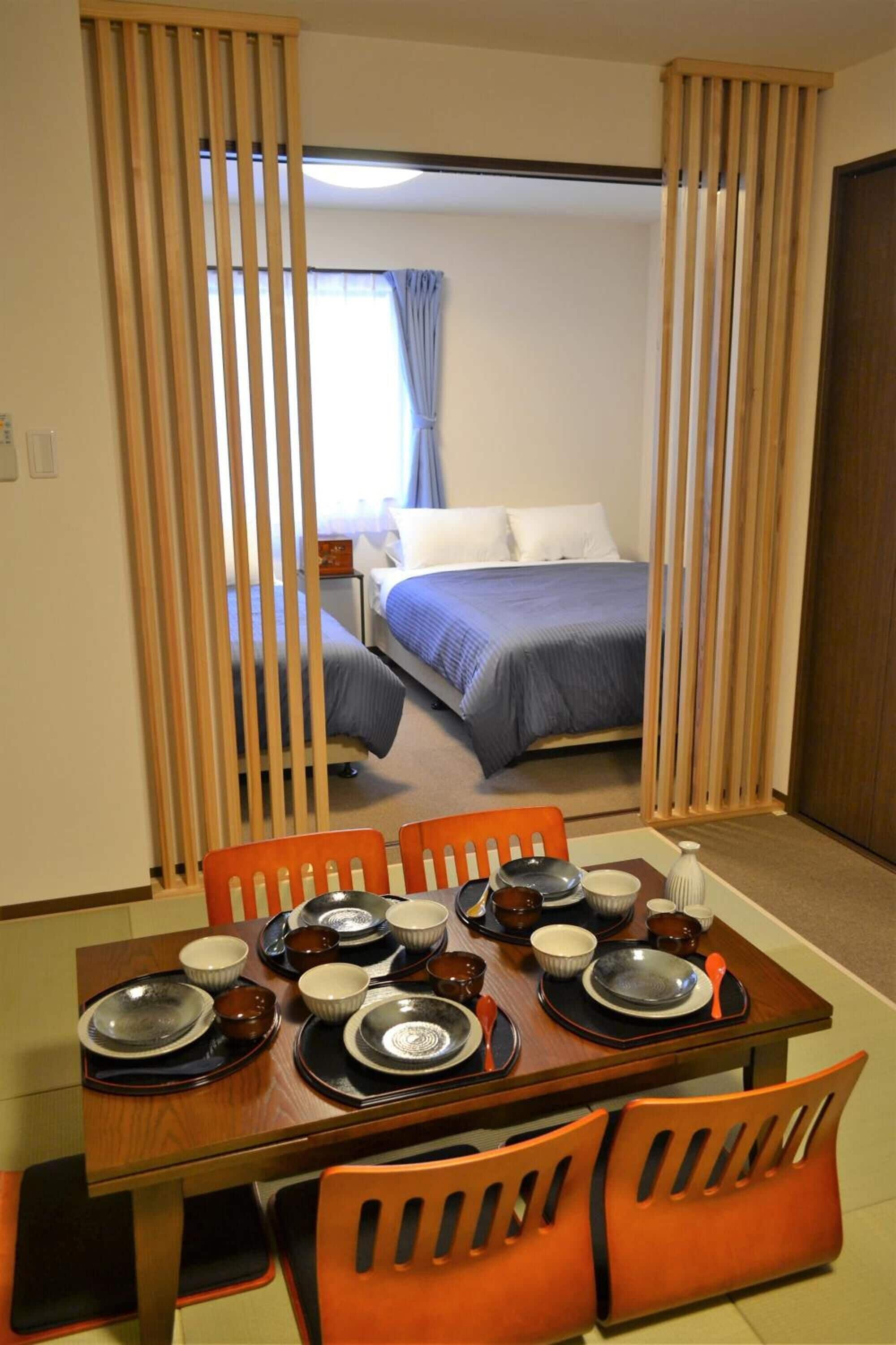 Une chambre d’hôtel avec une table à manger dressée pour quatre personnes, un lit avec une literie bleue et une porte coulissante à lames verticales.