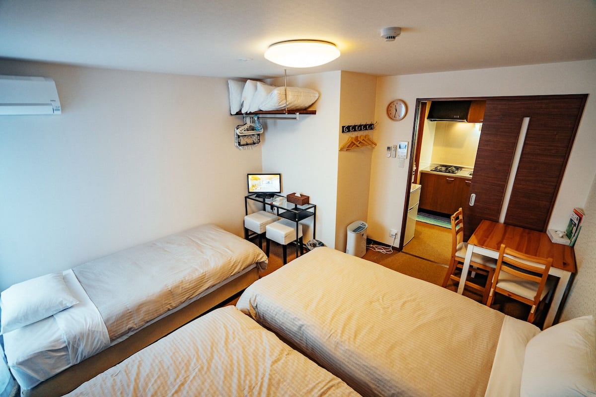 A compact hotel room with two single beds, a small desk with a computer, a chair, and a kitchenette area.